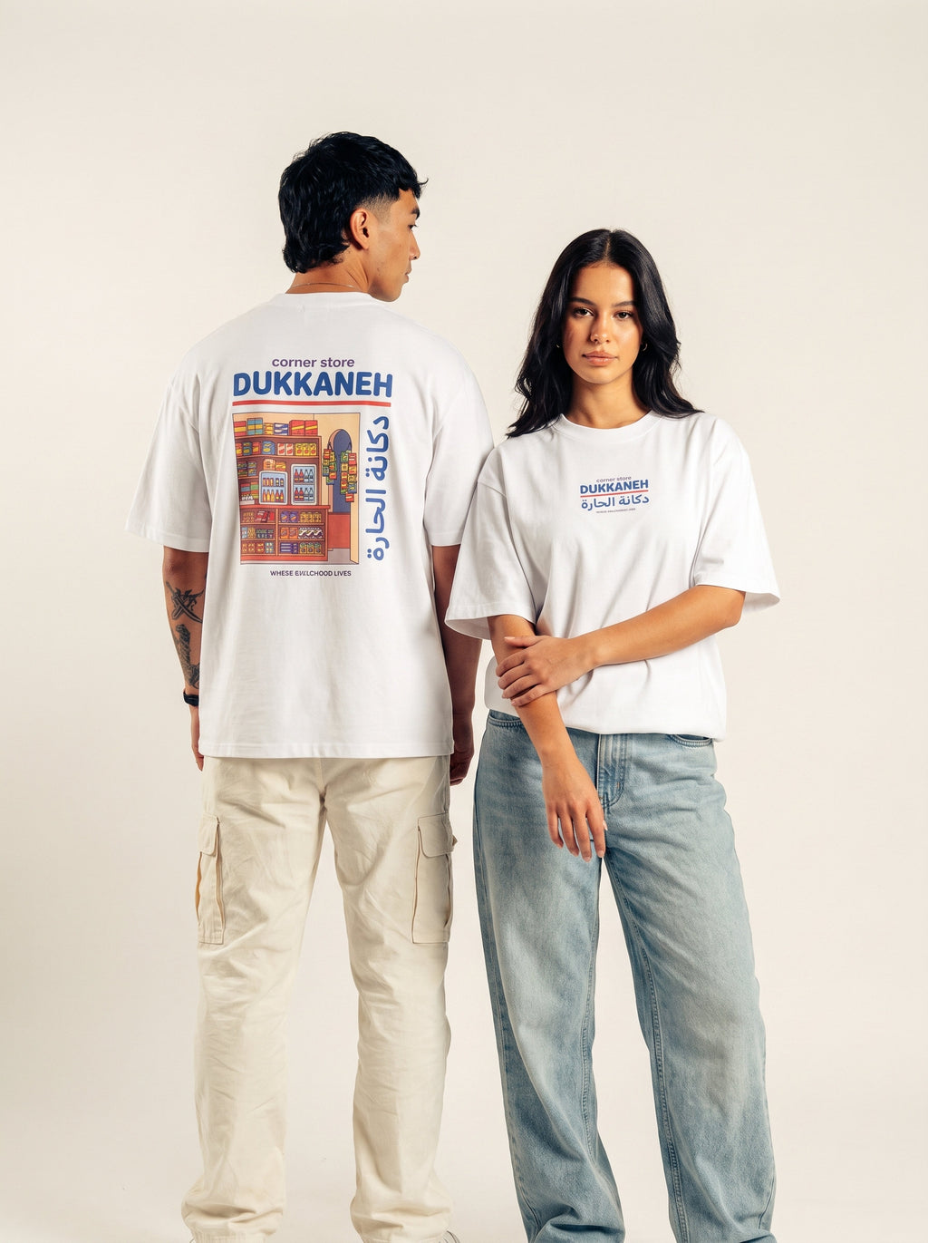 Two models showing the front and corner-store back graphic on a white Monani Dukkaneh T-shirt