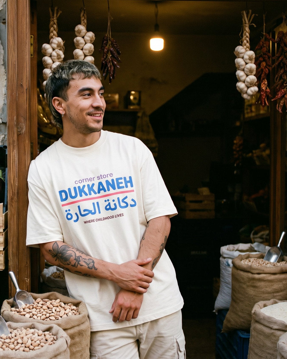 A man stands in front of a rustic Mediterranean-style shop doorway wearing a cream-colored t-shirt with blue and red text that reads 'DUKKANEH' and Arabic script. The shop is decorated with hanging garlic bulbs and dried peppers, and displays fresh produce including tomatoes, eggplants, and lemons alongside large burlap sacks of grains.